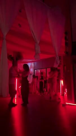 The scene is a dimly lit room with red lighting, where multiple couples are dancing. White sheets are draped from the ceiling, and neon lights are placed around the room. A fan is visible on the wall, and a screen displaying an image is set up in the background. People are mainly silhouettes, but some details such as clothing (dresses, t-shirts, shorts) are noticeable. The dancers are engaged in close contact and movement typical of partner dancing.