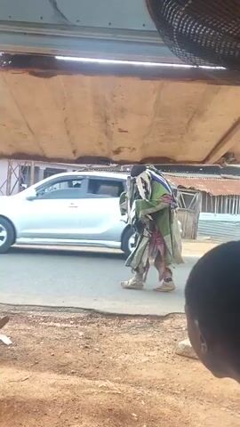From a slightly elevated indoor vantage point, we see a street scene. A person dressed in traditional attire, predominantly green and brown with white accents, walks across the street. The person is wearing a mask or head covering with strands hanging down. Behind them, people mill about on the street. A silver car is parked on the left side of the frame. A yellow auto rickshaw drives by on the street. The view is framed by a window or doorway with a metal awning above. The camera is focused on the person in traditional attire, following their movement. In the foreground, we see the back of two heads, one with dark hair and the other covered with a red cloth. The environment appears to be a bustling, possibly urban, area.