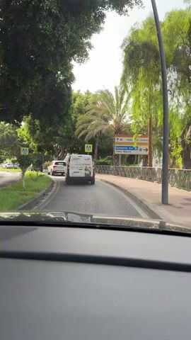 The video captures a view from inside a car driving on a road. The road is flanked by trees on both sides, with lush green foliage and palm trees visible. Pedestrian crossing signs are present on the side of the road. In front of the car, a white van with some lettering on its back is driving, followed by a white sedan. Further ahead, there are more cars and a green bus. Traffic signs indicating directions to "Valle San Lorenzo", "Autopista Sur", and "Cabo Blanco" are also visible. The weather appears to be daytime with a bright sky, suggesting it might be a sunny or partly cloudy day. The overall environment appears to be a suburban or semi-urban area with well-maintained roads and greenery.