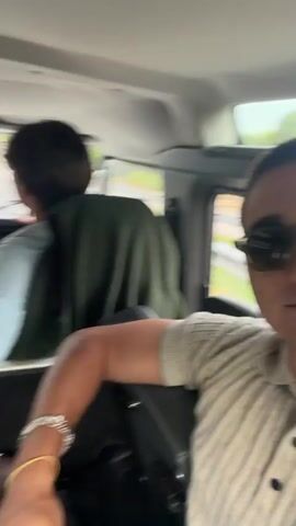 The video shows the interior of a vehicle traveling on a road. In the foreground, a person wearing sunglasses and a collared shirt smiles at the camera. In the middle ground, two people are visible in the front seats. The driver, wearing a dark shirt and a cap, is holding the steering wheel. The passenger next to the driver, wearing a light blue shirt and a dark green vest, is looking forward and gesturing with their right hand. In the background, the road and trees are visible through the windshield. Other passengers are partially visible in the back seats. The overall atmosphere appears casual and friendly.