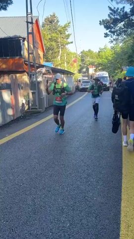 A group of runners are participating in a race on a paved road. The runners are wearing athletic clothing and bib numbers. Some runners are wearing hydration packs. An ambulance is visible in the background, and there are trees and buildings on either side of the road. The sun is shining, indicating it is daytime. The runners are moving in the same direction.