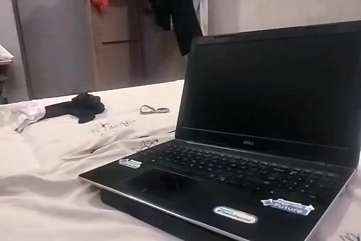 A laptop with a Dell logo is open and placed on a black cooling pad on a bed. The laptop has stickers on its surface. In the background, a closet with clothes hanging on it is visible. There are also a pair of glasses and a black object on the bed. The lighting is dim.