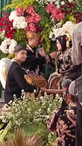 A wedding ceremony is taking place, with a bride and groom seated on a decorative couch surrounded by abundant floral arrangements. The groom, wearing traditional Javanese attire including a batik sarong and peci (cap), is in the process of drinking from a glass. The bride, also in traditional Javanese wedding attire, which includes a black dress with intricate gold embroidery, a headscarf, and a bridal headdress, is receiving a drink from him. Another young man, dressed in a similar black outfit and peci, sits to the left, offering a tray with what appears to be more drinks. A person with a camera is visible in the foreground, capturing the moment. The setting is outdoors, with a large green backdrop behind the floral decorations.
