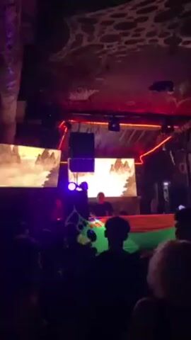 The video shows a club with a DJ playing music for a crowd. The DJ is standing behind a colorful booth, with two screens displaying visuals.  The crowd is mostly in silhouette, dancing in the dimly lit space. The ceiling has a unique pattern with holes, and the lighting is mostly purple and red. There's also a pipe running along the side of the ceiling.