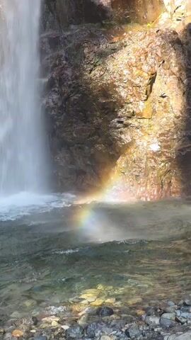 A slow-motion video shows a waterfall cascading down a rocky cliff face. The water crashes into a pool below, creating a mist that refracts sunlight to form a small rainbow. The rocks are various shades of brown and orange, and the water is a murky green. The bottom of the pool is visible, revealing a bed of smooth, gray pebbles. The overall scene is tranquil and serene.