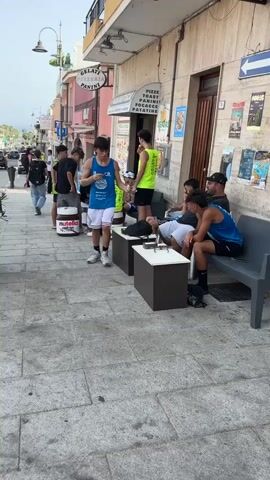 The video pans across a street scene, capturing an outdoor area with several young males. In the foreground, a young man wearing a blue athletic jersey and white shorts is standing, holding something to his mouth, possibly eating or drinking. To his right, another young man in a bright green jersey is seated, looking at his phone. Two other young men are seated on a bench to the right, also looking at their phones. A large round object with "Nutella" branding is visible near the center of the frame. In the background, there are buildings, including one with a sign that reads "GELATERIA PIZZERIA PANINI." Cars are parked and driving on the street. The ground is paved with cobblestones and there are planters with greenery. The overall atmosphere appears casual, possibly a town square or street cafe. The video also briefly shows a red van driving away.