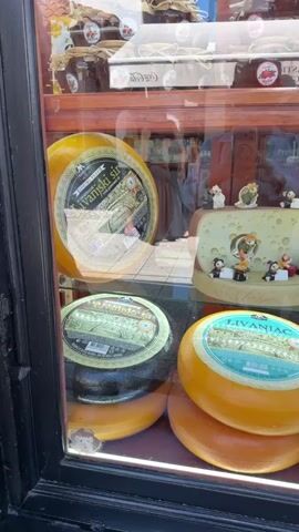 The video shows a close-up view of a cheese display in a shop window. The window features several large, round cheeses stacked on top of each other. The cheeses are of different colors, ranging from yellow to dark brown. Some have labels with text that appears to be in a Slavic language, with one label clearly visible reading "LIVANJAC".

Behind the cheeses, there is a decorative display of a cheese block with small figurines, possibly mice or other characters, interacting with it. Further back, more cheeses are visible, including one with a label that reads "100% Kozji sir", indicating it is goat cheese. Above the cheese displays, shelves are stocked with jars of what appear to be jams or preserves, also with labels.

The scene is set indoors, within a shop, with reflections of the outside visible in the glass. Through the reflections, glimpses of people and buildings can be seen, suggesting an urban environment. The lighting indicates it's daytime. The overall impression is of a well-stocked food shop, likely specializing in dairy products, with an emphasis on presenting the items in an attractive way. The small figurines on the cheese display add a whimsical touch.