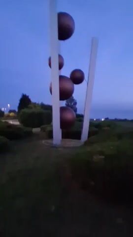 A vertical sculpture with four spherical elements is visible against a twilight sky. The sculpture consists of two white vertical beams supporting the spheres, which are dark and appear metallic. The spheres are arranged in a stacked, slightly staggered formation. Surrounding the sculpture is a low-lying landscape with manicured bushes and grass. The overall lighting suggests it is either dawn or dusk.