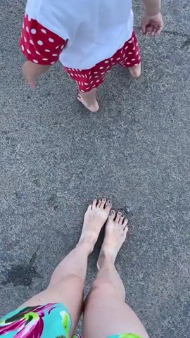 The video shows a high-angle view of two pairs of bare feet walking on a textured, grey, asphalt surface. In the background, a young child is walking away from the camera. The child is wearing a white t-shirt with a red polka dot pattern on the sleeves and shorts, also with red polka dots. The child's hair is pulled back in two small pigtails. The person filming appears to be walking behind the child. The lighting suggests it is daytime, possibly late afternoon or early evening, as there are distinct shadows cast on the ground. The location is identified as Сям-Можга, Russia. There is no visible text in the video. The scene evokes a sense of casual outdoor activity, possibly a walk in a park or along a street.
