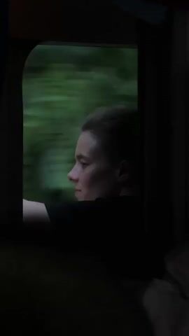 This media file shows a close-up profile view of a young person with short, pulled-back hair, looking out of a train window. The person is wearing a dark t-shirt and their left arm is resting on the windowsill. The window frames a blurred green landscape, indicating the train is moving at speed through a natural setting. The interior of the train carriage is dark, with the light primarily coming from the window, creating a silhouette effect for the person and the immediate surroundings. The lighting suggests it could be late afternoon or early evening, or a heavily overcast day. The overall mood is contemplative and perhaps a little melancholic, with the blurred motion emphasizing a journey or transition. There is no visible text in the media file. The scene is set within a train moving through an area that can be contextually placed in Rakovica Urban Municipality, Serbia.