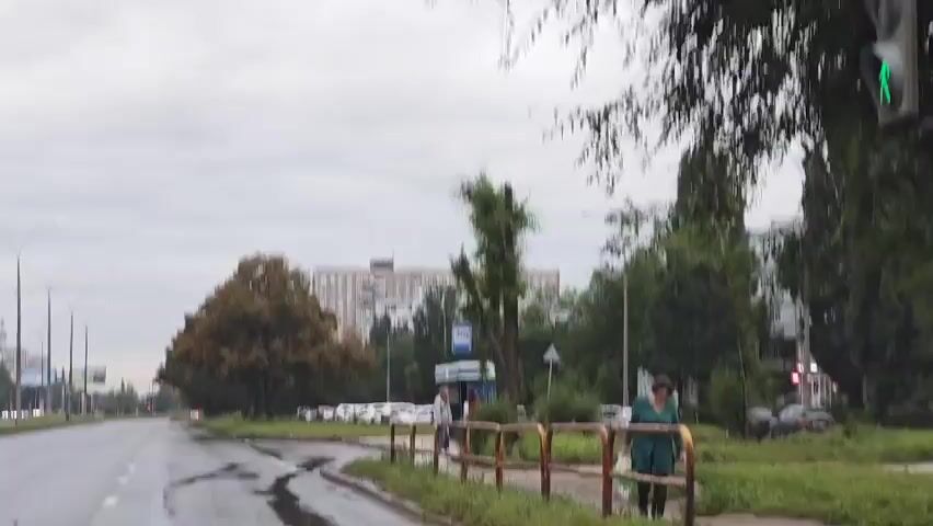 A man wearing a black baseball cap and a red and black jacket is walking across a wet crosswalk. He is holding a white bottle in his left hand. Two other people are walking behind him, and a woman in a green coat is standing on the sidewalk to the right of the crosswalk. The road is wet and reflects the overcast sky. Trees and buildings are visible in the background.