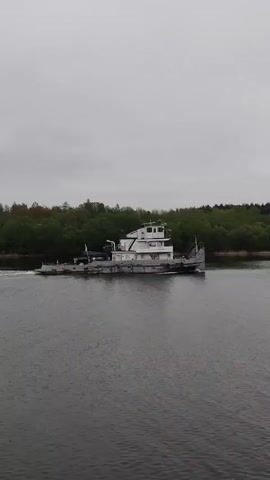 The video shows a white and gray tugboat moving along a river under a cloudy sky. The boat is traveling from left to right in the frame, creating a small wake in the water. Lush green trees line the riverbank on both sides. The water is dark gray. The overall scene appears calm and overcast.
