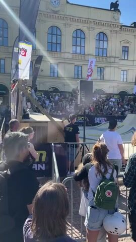 A sunny day event features a skateboarding competition in a public square in front of a large, ornate building with multiple windows. A crowd of spectators watches from behind a metal fence. A skateboarder performs a trick on a ramp. There are banners and speakers visible around the event area.