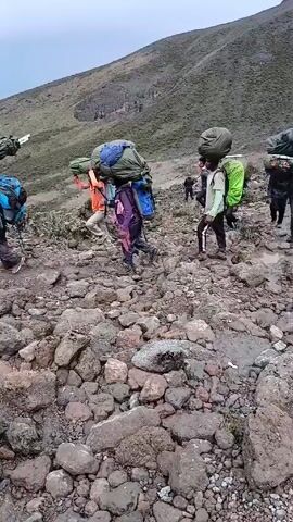 Several people are walking across a rocky landscape. Each person is carrying a large duffel bag on their head in addition to a backpack. The bags appear to be green. The landscape is covered in rocks, with a hillside in the background. The sky is overcast.