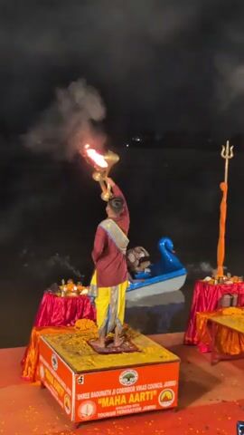 A person stands on a platform holding a flaming torch aloft. They are dressed in a maroon tunic and a yellow dhoti. The platform has red coverings and is decorated with yellow flowers. In the background, a boat with two people is on the water. Two tables draped in red fabric with offerings are visible on either side of the platform. Smoke billows from the torch. The scene appears to be an evening ritual or ceremony.