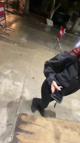 The video begins with a blurry shot, then refocuses to show four individuals standing on a sidewalk at night. One person, wearing a black hoodie and a cross-body bag, is closest to the camera and smoking a cigarette. A second person, wearing a gray hoodie, points away from the camera. A third person is wearing a black baseball cap and a black jacket. The fourth person is wearing a black beret and a black jacket and holding a phone. The sidewalk is concrete, and there are buildings and trees in the background. The scene is lit by streetlights.