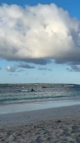 The video shows a beach scene with white sand in the foreground. The ocean is visible with waves rolling onto the shore. Several people are in the water, some on what appear to be surfboards or paddleboards. There are also two jet skis in the distance. The sky is blue with large, fluffy white clouds. The horizon shows a line of trees or land in the far distance. The water is a clear turquoise color. The lighting suggests it is daytime, likely afternoon or early evening, given the soft light and the contrast of the clouds.