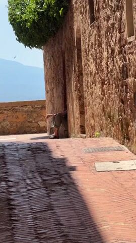 A person is sitting in a recessed doorway of a stone building. They are wearing green pants and dark shoes. The person is leaning against the wall and appears to be relaxed. The doorway is partially obscured by ivy. The ground in front of the doorway is paved with cobblestones and has a textured pattern. In the background, there is a wall and a distant mountain range. A bird is flying in the upper left corner of the frame. The sun is casting shadows on the ground.