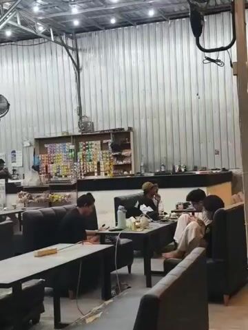 The video shows the interior of a cafe or restaurant. There are several tables with seating, and a counter area in the background where food and drinks are likely prepared or sold. Various people are seated at the tables, some interacting with each other, while others appear to be looking at their phones or working. The environment is lit by overhead lights and appears to be indoors. There are shelves stocked with items behind the counter. A fan is mounted on the wall. The overall atmosphere seems relaxed and casual.