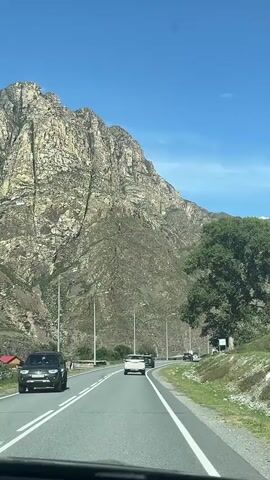 The video shows a view from inside a car driving on a paved road. The road has two lanes in each direction, separated by white lines. Several cars are visible on the road ahead. To the left of the road is a large, steep, rocky mountain with patches of greenery. To the right of the road is a grassy embankment with a large tree. The sky is clear and blue. The video appears to have been filmed during the day.