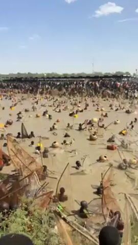 The video shows a large group of people participating in a fishing event in a shallow, muddy body of water. The people are mostly submerged, with only their heads and shoulders visible. Many are using fishing nets or baskets to catch fish. There are various floating objects scattered in the water, including gourds and plastic containers. A large crowd of spectators is visible along the shore, watching the event. The sky is partly cloudy and the overall environment appears to be outdoors in a rural or undeveloped area.