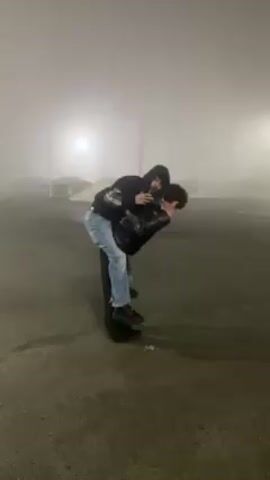 In a foggy outdoor environment, two individuals engage in a playful activity. One person, dressed in light blue jeans, a black hoodie, and a black leather jacket, is perched on the back of the other. The person on whose back the other is standing is wearing all black, and they are moving around with the person on their back. The person being carried is looking at their phone. In the background, several cars and a pile of sand are visible through the fog. Street lights cast a faint glow. The ground appears to be a parking lot or paved area.