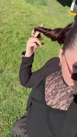 The video shows a person in a black outfit standing on a green lawn. The person is holding the end of their dark brown ponytail and appears to be twisting it. They are wearing sunglasses and a black jacket over a black top with lace detailing. The background is a sunny green lawn. Other people are vaguely visible in the background. The camera appears to be hand-held and at a high angle, looking down at the subject.
