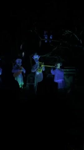The video shows a dark, spooky scene. Four ghostly figures are visible, each playing a different instrument. The figures appear to be translucent and are illuminated with blue and green light. The first figure on the left is holding a banjo-like instrument. The second figure from the left is playing a flute. The third figure from the left is playing a set of bagpipes. The fourth figure on the right is playing a trumpet. The background is dark and appears to be a graveyard with tombstones. There are also some trees visible in the background. The overall atmosphere is eerie and unsettling.