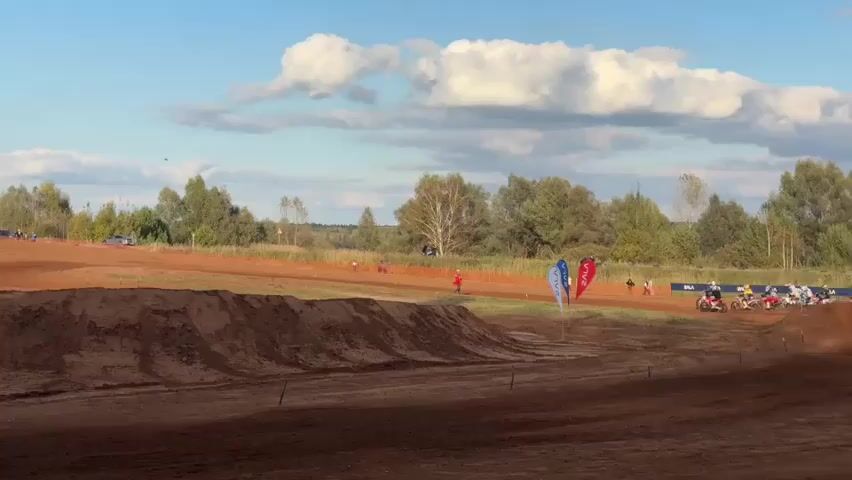 A motocross race is in progress on a dirt track. Several riders on motorcycles are visible, racing around a curve. The track is lined with trees and foliage, and a blue sky with scattered clouds forms the backdrop. There are also flags and some spectators visible in the distance. The foreground is dominated by a large dirt jump.