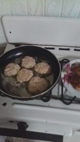 The video shows a stovetop with a frying pan containing several uncooked patties of food, likely meat or vegetable-based. The patties are light in color and appear to be simmering in oil or fat. Above the pan, on a rack, sits a plate with a few cooked patties of similar appearance. The stove is white and appears somewhat older, with some visible wear. The surrounding environment is a kitchen, indicated by the stove and possibly tiled walls in the background. No people are visible in the frame.