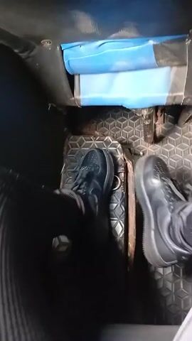 The video shows someone in a vehicle with their feet on the floor. Both feet are wearing black sneakers with white soles. The left leg is covered in black fabric and the right leg in gray striped fabric. The vehicle floor has a textured metal plate design. The upper part of the frame includes a partially visible blue object. The video is close-up and the camera is moving slightly.