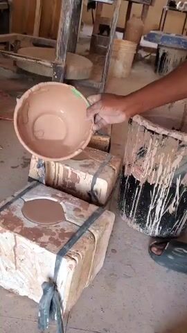 Here is a description of the video:

The video shows a person’s hand holding a brown bowl in a workspace. The bowl is empty except for a thin layer of brown clay residue. Below the hand are two stacked, light-colored blocks, bound together with black straps. Each block has a circular depression filled with liquid brown clay. To the right of the blocks, there is a paint-splattered black and white bucket, and the person is wearing a slide sandal. 

In the background, there's a cluttered workspace. There are wooden tables and various containers, some of which appear to have been used for mixing or storing clay. The light is bright, suggesting the scene is indoors with natural light entering the space.