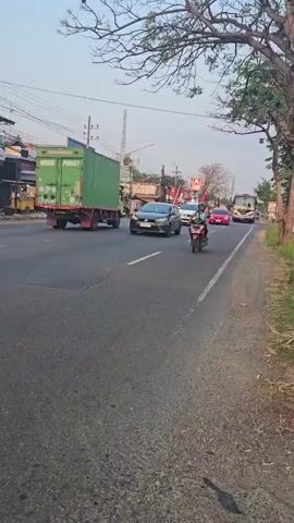 A low-angle, eye-level shot follows a motorcycle as it travels down a paved road. The motorcycle has a rider wearing a helmet and a teal top. The rider is approaching an intersection where several cars and a bus are present. There are buildings and trees lining the road. The sky is clear and the lighting suggests it is either sunrise or sunset.