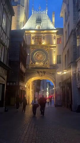 A nighttime street scene features a prominent ornate clock tower archway in a European-style city. The clock tower, illuminated by spotlights, has a grand gold-colored clock face with intricate designs. Below the clock, an archway opens to a bustling street with additional lighting. People walk along the cobblestone street, some in pairs, others individually. Buildings on either side of the archway are also lit, revealing architectural details such as timber framing and multiple stories. The sky above is a dark blue, indicating evening or early morning. The atmosphere is vibrant, with the sounds of people talking and general urban ambient noise.