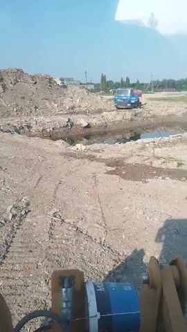 A construction site with a blue truck with an orange tank is visible in the background. In the foreground, a piece of construction equipment is partially visible. The ground is rough and appears to be dirt with some debris. There are mounds of dirt and a small body of water on the site. The sky is blue with some clouds.
