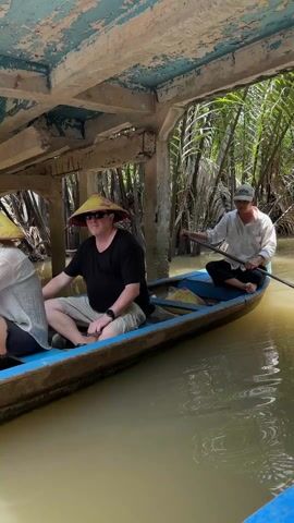 A man is seen on a blue boat as he uses a long paddle to propel himself away from under a concrete building. The boat moves forward into a narrow canal lined with tall green palm trees. The water is murky brown in color.
