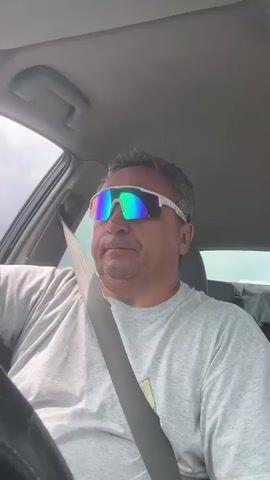 A man is driving a car. He is wearing a gray t-shirt and white sunglasses with mirrored lenses. He has short, gray hair and is looking forward. The seatbelt is visible across his chest. The interior of the car is visible, including the ceiling and part of the dashboard. The lighting suggests it is daytime.