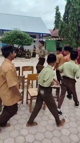 A group of young people in school uniforms are playing a game of musical chairs outdoors on a paved area. There are several chairs arranged in a circle. The people are walking or running around the chairs. The environment appears to be a schoolyard with buildings and trees in the background. Some of the people are wearing tan shirts and brown pants, while others are wearing white shirts with brown pants. Some are also wearing black caps. The sky is overcast.