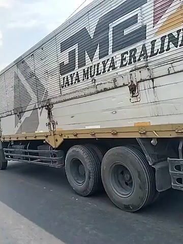 A truck with the words "Jaya Mulya" on its side is driving on a paved road. The truck is a light brown color, and its cargo hold is silver. The road has a barrier painted in black and yellow. In the background, there are buildings and streetlights. The sky is cloudy. The camera is moving with the truck.