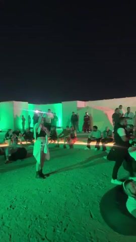 The video shows a vibrant outdoor gathering at night. People are scattered across the sand, some dancing to music, others sitting and relaxing on beanbag chairs. In the background, a large architectural structure is illuminated with green lights. There's a food stand on one side, and many people are lined up there. The scene is active and appears to be a lively social event.