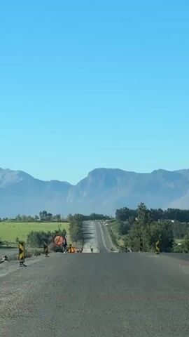 The video shows a straight asphalt road stretching towards a mountain range under a clear blue sky. The road has a slight incline and is marked with yellow and black chevron signs on either side. There is a construction zone with cones and signs indicating no bicycles allowed, and workers are present. The landscape consists of green fields and trees, creating a rural scene.