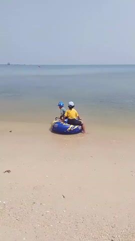 Two children, one wearing a yellow shirt and the other a blue hat, sit on a blue inflatable tube at the edge of a sandy beach. The ocean stretches out behind them under a clear, bright sky.