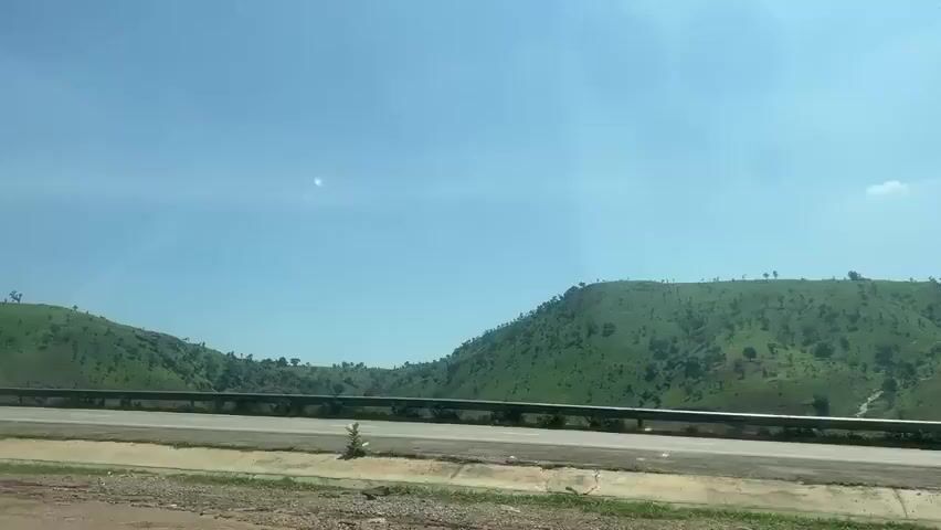 The video captures a scenic drive along a highway with lush green hills on both sides. The sky is clear and blue, indicating good weather. A car is visible on the left side of the road, suggesting movement. The view through the car window shows reflections and glare, hinting that the footage is taken from inside a moving vehicle. The focus is on the expansive landscape, with no specific human activity or interaction highlighted.