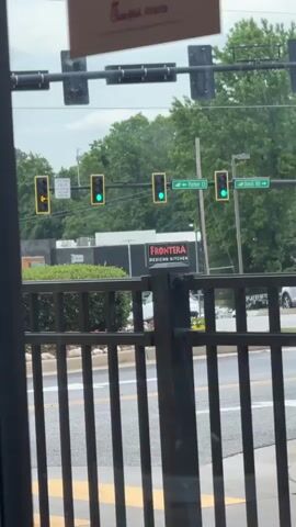 The view from indoors looks out at a street. A black metal fence partially obscures the view. Above the fence are green traffic lights for each direction. The traffic lights turn yellow. A sign for Frontera Mexican Kitchen is also in view. Trees are in the background. A car drives by.