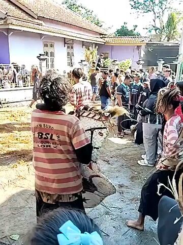 The video shows a gathering of people in an outdoor space, likely a yard or street, in front of a pale purple house with a brown roof. The ground is covered with loose straw or dry grass. There are multiple people, mostly men, standing and moving around. Several individuals are dressed in patterned shirts and black pants, and some carry what appear to be drums with star symbols. Three figures dressed in black outfits with large, dark, furry head coverings are the center of attention. There is a thin pole visible, extending upwards into the sky. Some people in the background are standing behind a low fence or barrier. The overall atmosphere seems festive or ceremonial.