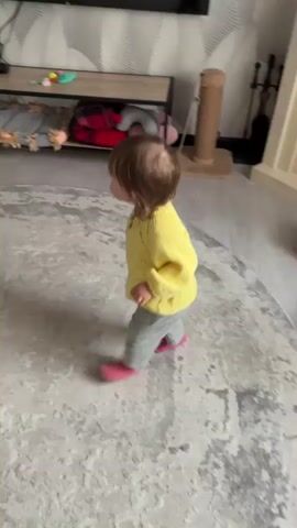 A young child wearing a yellow sweater and grey pants walks across a rug in a room. The child's back is to the camera. The room has patterned wallpaper, a shelf unit with objects, a television, and a stuffed dog. The child walks towards a doorway and then turns.