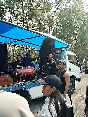 The video begins with a close-up shot of a truck bed filled with musical equipment and people. A blue tarp is stretched over a frame, creating a makeshift canopy. On the truck, there are drums, speakers, and a generator. Several men are seated or standing on the truck, some appearing to be preparing or operating the equipment. A woman with a black cap is visible in the foreground, facing away from the camera. The scene appears to be outdoors, with trees and foliage visible in the background. The video then pans out to show a group of people gathered on a dirt road lined with trees. Some people are walking down the road, while others are standing and observing. The truck with the musical equipment is positioned on the side of the road. The overall atmosphere seems to be that of an outdoor event or gathering.