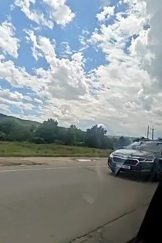A time-lapse video shows a view from a moving vehicle of a rural landscape under a partly cloudy sky. A paved road is in the foreground. Beyond the road is a grassy area with scattered trees and bushes. In the distance, rolling hills are covered with vegetation. A power pole with wires is visible on the right side of the frame. The clouds are white and puffy, with some appearing larger and more defined than others. The sky is a clear blue. The time-lapse effect makes the clouds appear to move and change shape rapidly. There are no people or animals visible in the video.