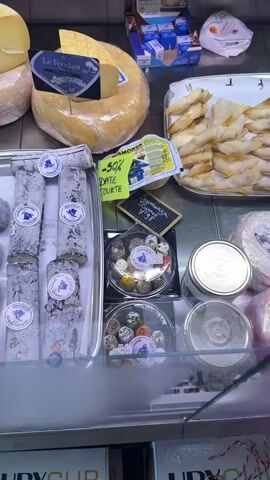 The video pans across a display case filled with various cheeses and other food items. In the foreground, there are trays of small, round cheeses, some coated in herbs or spices, and others plain white. Behind these, there are long, log-shaped cheeses, some covered in a blue or grey mold. Larger wheels of cheese are visible in the background, one of which has been cut to reveal its interior. There are also some packaged items, including what appear to be phyllo pastry rolls and small containers of what could be quail eggs or chocolates. Several price tags and labels are attached to the products. The lighting suggests an indoor environment, likely a cheese shop or market. No people are visible in the video.