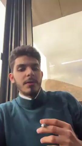 A young man is shown in a close-up shot, positioned indoors near a large window. He has dark hair, a mustache, and a goatee. He is wearing a dark teal sweater over a white collared shirt. The man is smoking a cigarette, and the exhaled smoke is visible rising around his head. The window behind him offers a view of a landscape featuring a hill or mountain range under a cloudy sky. The setting appears to be a room with vertical blinds and a plain interior.